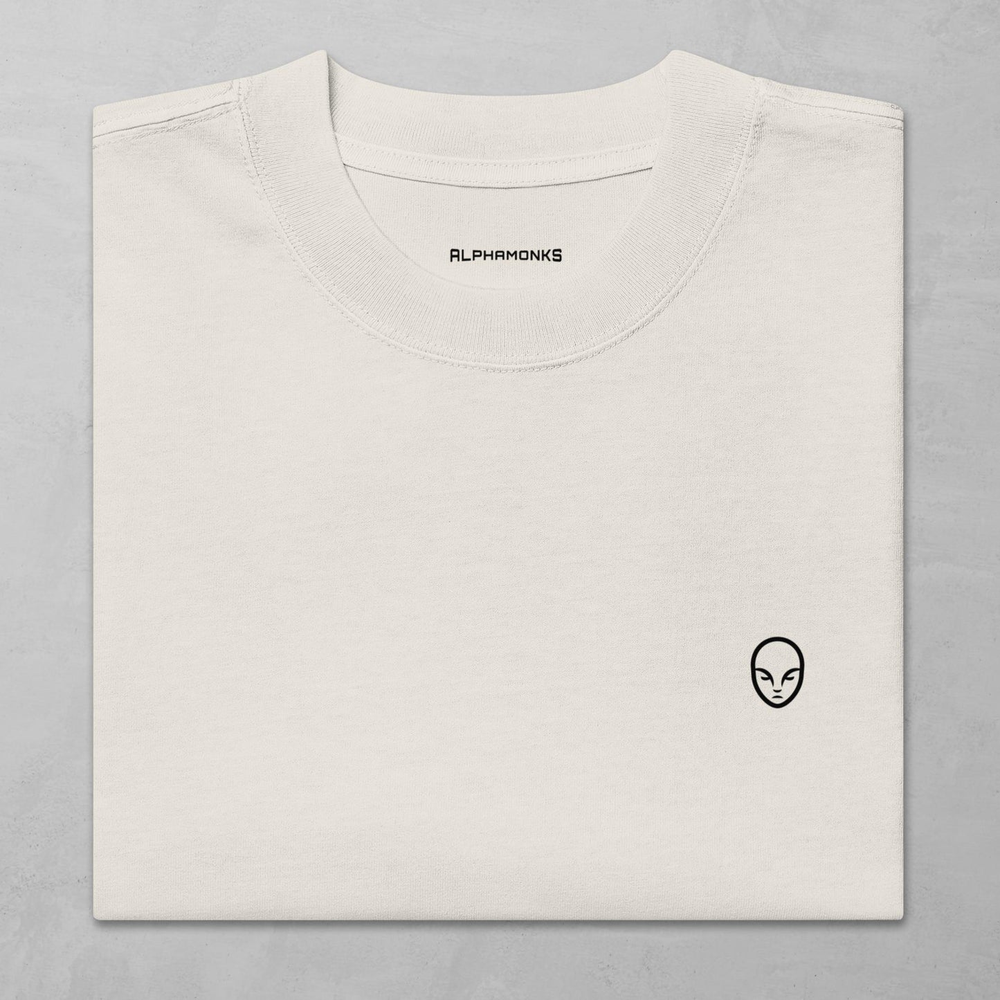 Close-up of a folded faced bone oversized t-shirt with a minimalist Alphamonks logo design on the left chest and 'Alphamonks' branding inside the collar