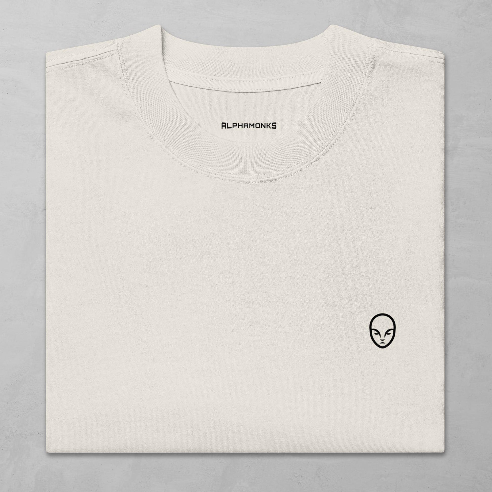 Close-up of a folded faced bone oversized t-shirt with a minimalist Alphamonks logo design on the left chest and 'Alphamonks' branding inside the collar